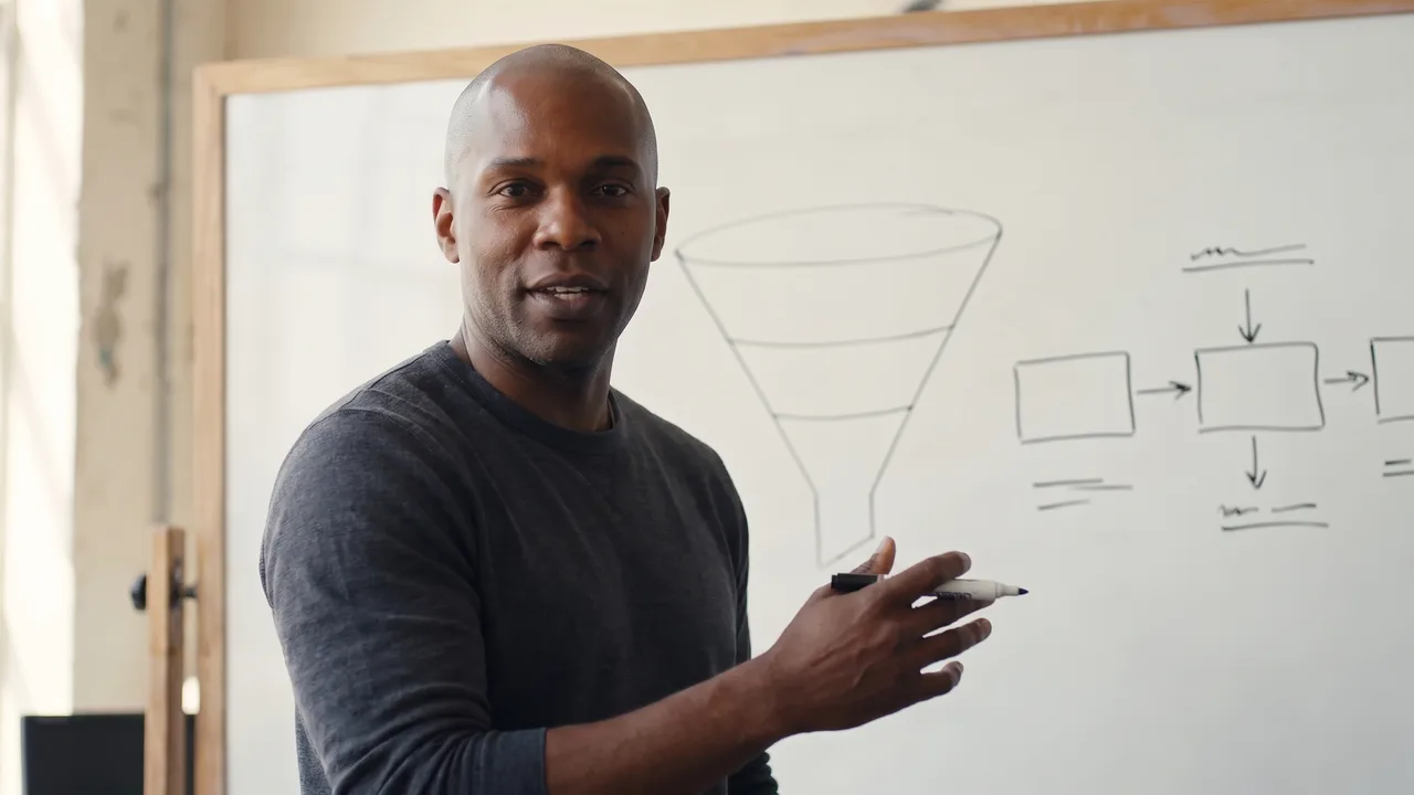 A sales coach beside a whiteboard with a funnel drawn on it, teaching to the camera.
